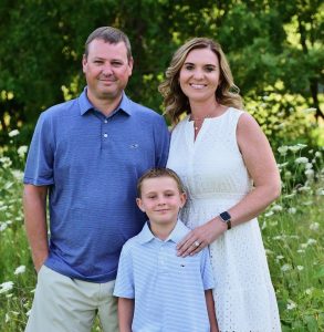 photo of the Denman family. Man wearing a blue shirt and shorts stands next to a woman with short curly hair in a white dress. She has her hand on the shoulder of their young son who is wearing a light blue polo shirt. 